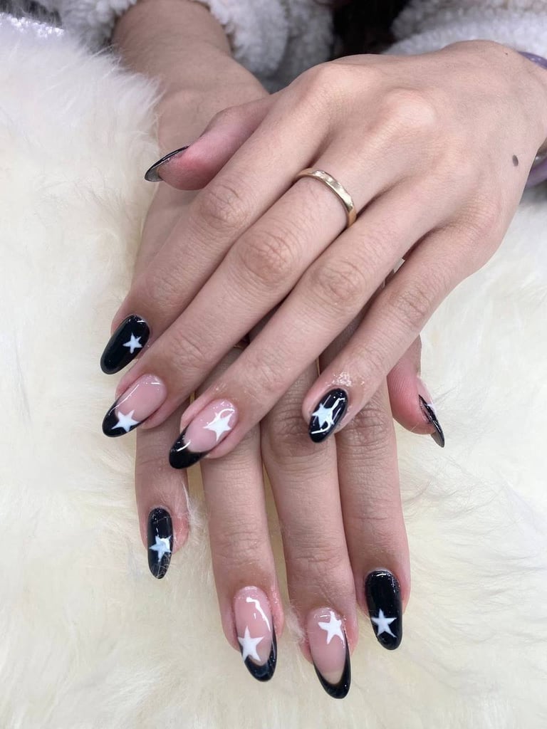 Close-up of manicured nails with black and pink design, white stars and lines, gold band ring on white textured background