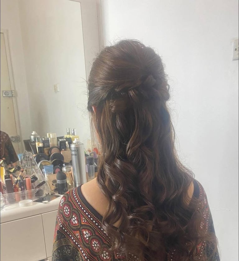 Woman with long wavy brown hair styled in a half-up style, wearing a patterned burgundy top, photographed from behind at a vanity mirror